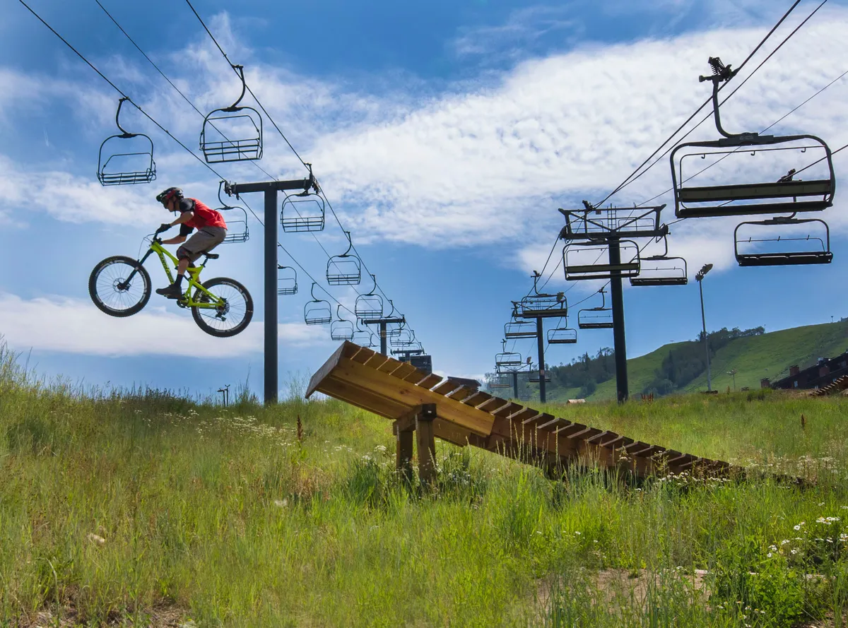 A dynamic exterior scene showing a mountain biker on a ramp near a ski lift, set against a lush, green backdrop. The area hints at access to both summer and winter recreation, capturing the spirit of adventure and proximity to trails described in the section.