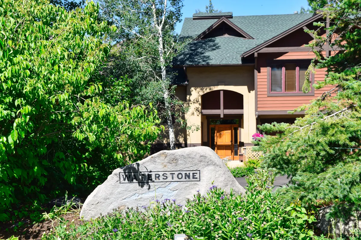 A welcoming view of the property entrance, with a large rock featuring the 'WATERSTONE' inscription set among lush greenery and aspen trees. The entrance is surrounded by well-maintained landscaping and hints at nearby nature trails, inviting guests to explore the area.