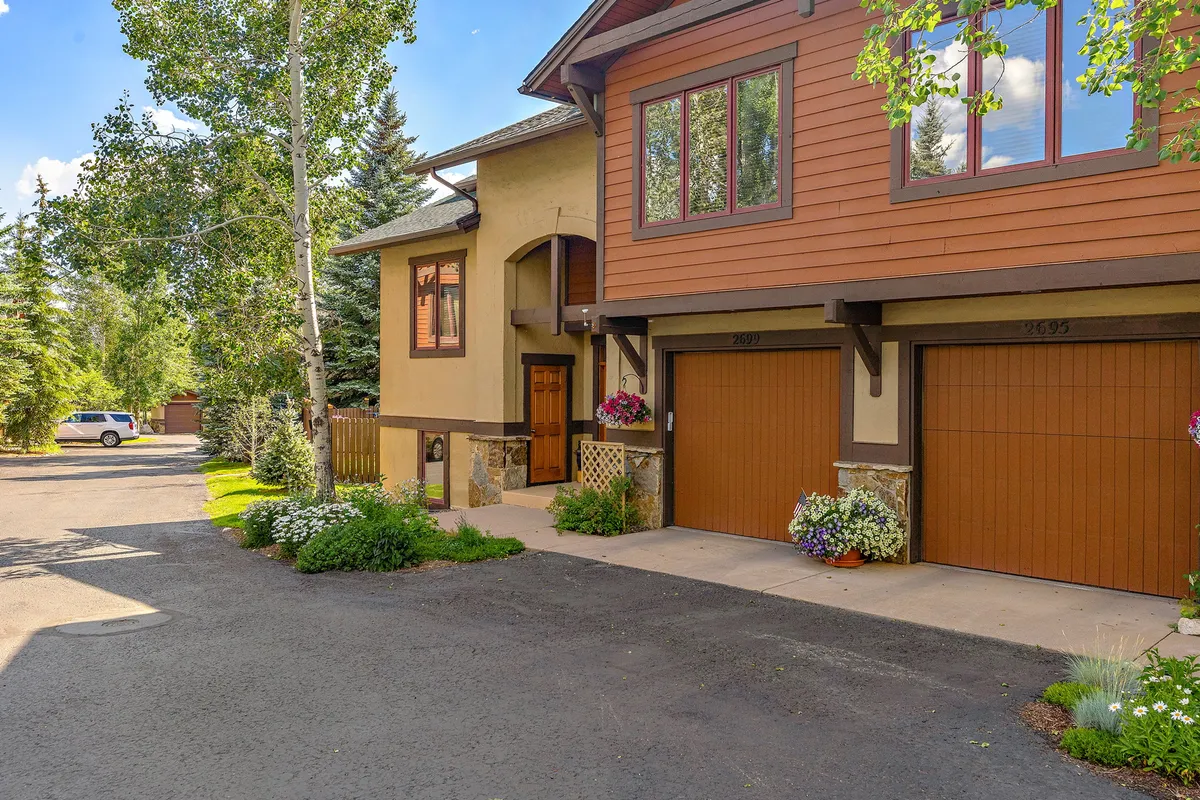 The image shows the front entrance of a townhome, framed by garage doors, flower planters, and windows. The setting suggests a welcoming, private home within a friendly community, ideal for relaxing and socializing.