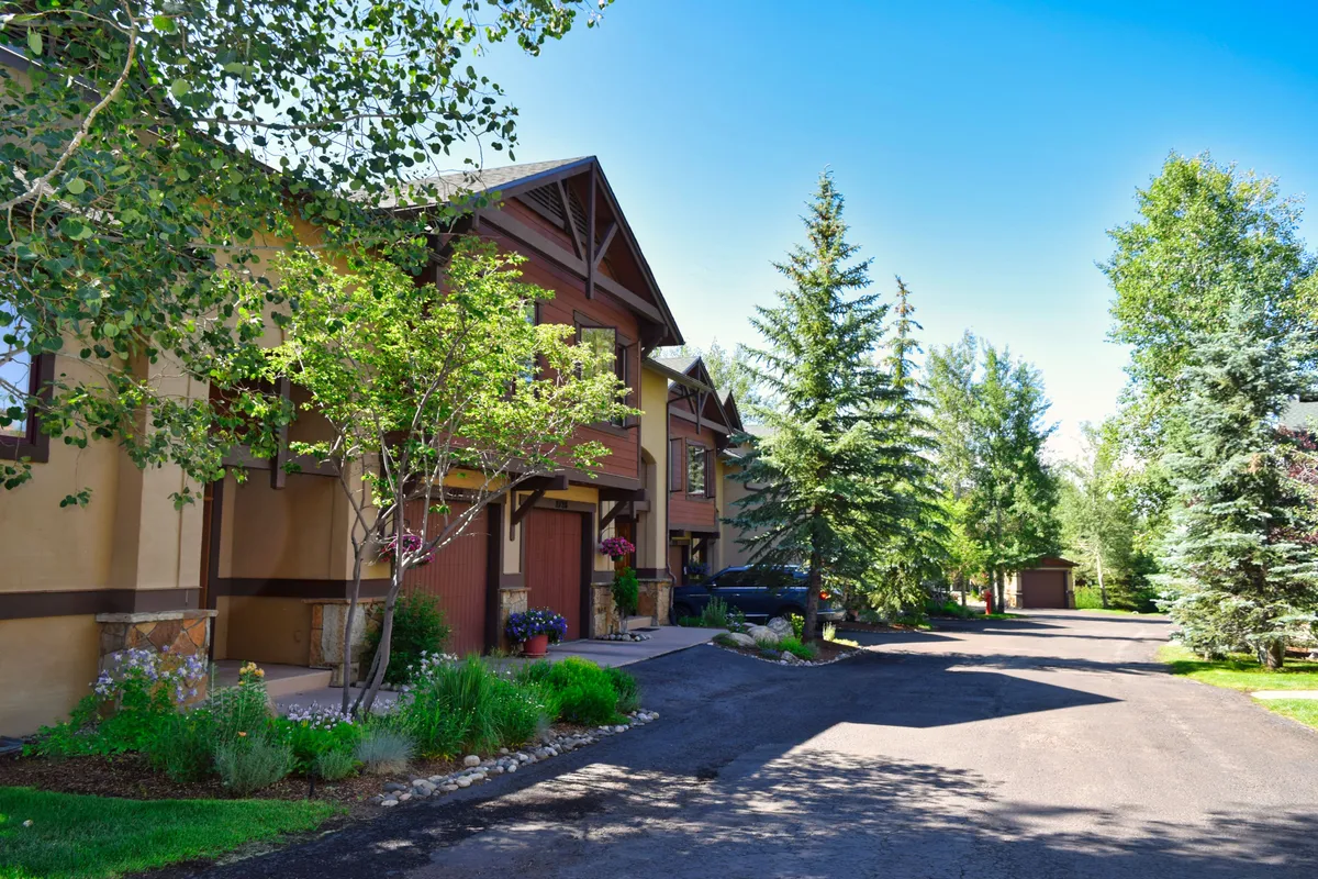 The front facade of the homes is bordered by lush landscaping and a well-maintained pathway, with sunlight filtering through the trees. The scene suggests easy access to walking and biking routes right from the doorstep, as well as opportunities to socialize in garden spaces.