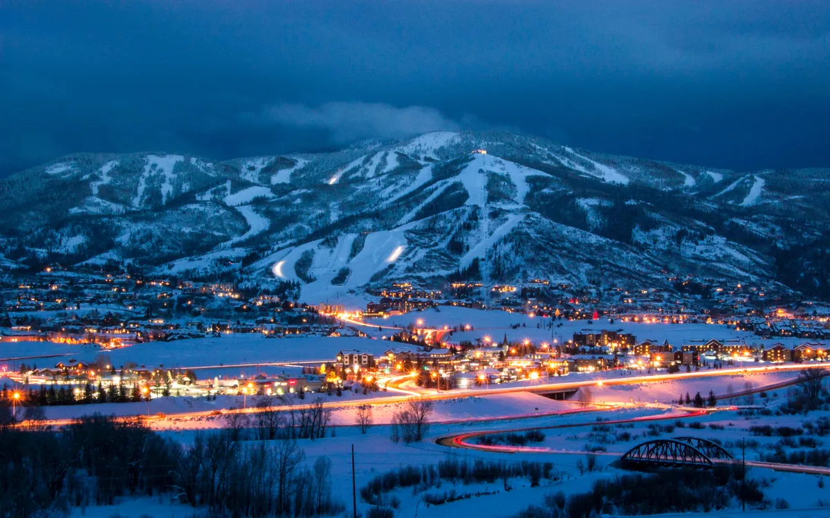A panoramic view of snow-capped mountains overlooking Steamboat Springs, with the town illuminated in the evening and a winding road leading into the heart of the resort. The scene captures the enchanting atmosphere of a winter getaway, blending natural beauty with vibrant community life.
