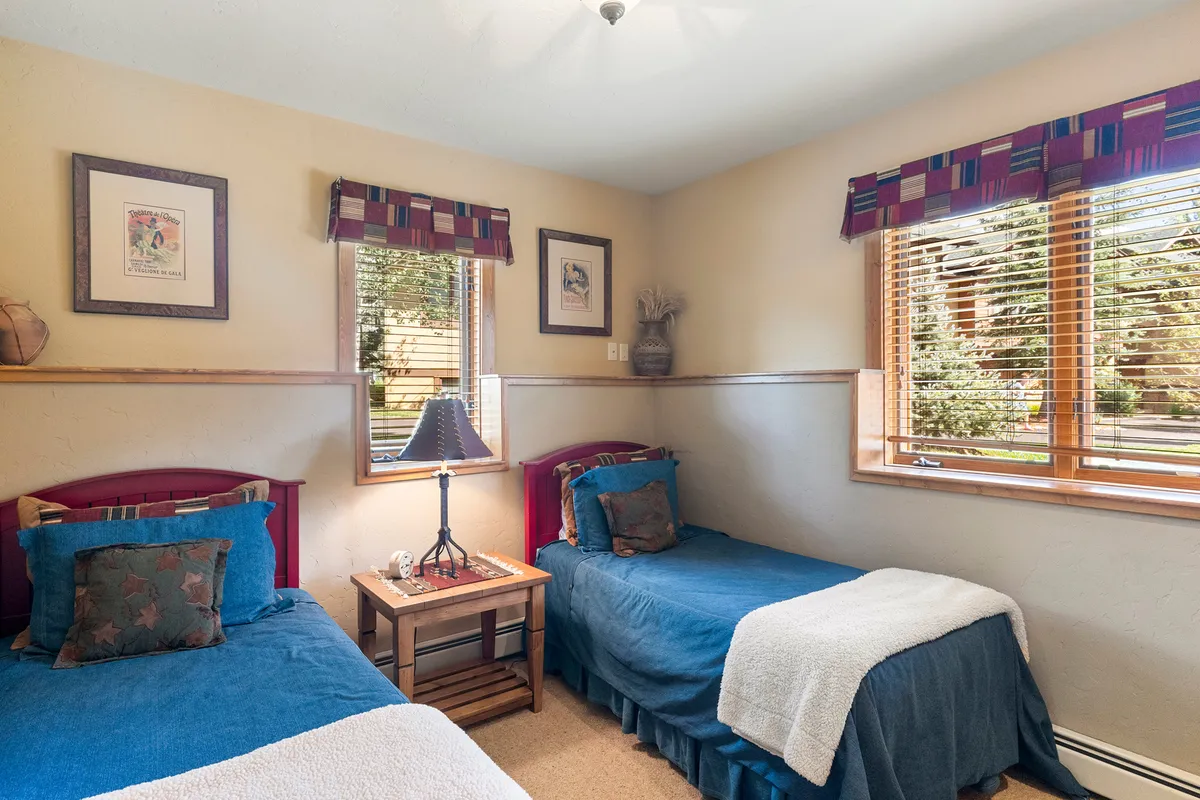 A cozy bedroom with two twin beds dressed in blue covers, a central nightstand with a lamp, framed artwork, and a large window offering a view of lush greenery outside. The room is bathed in gentle, natural light, creating an inviting and family-friendly atmosphere.