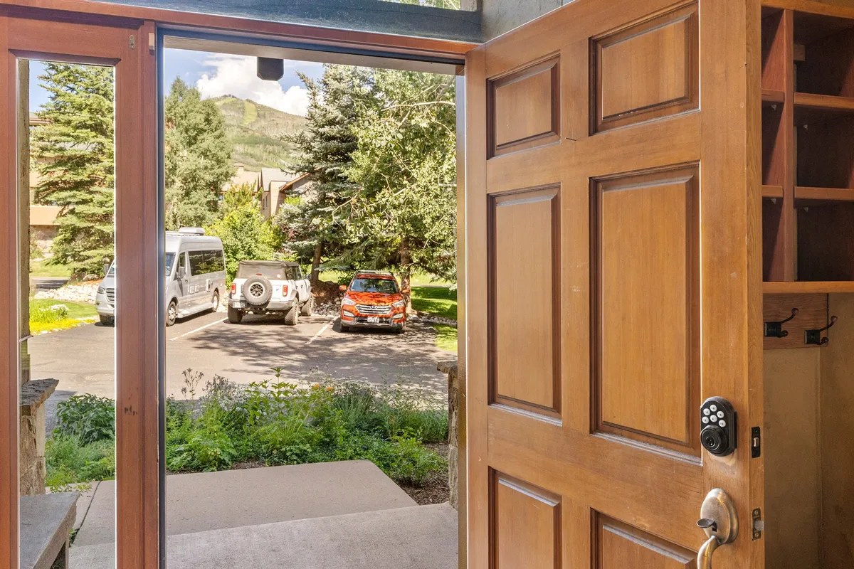 This image highlights a wooden front door opening onto a landscaped garden with parked vehicles, blending the transition between private home and the outdoors. The scene suggests arrival, relaxation, and connection to the natural surroundings.