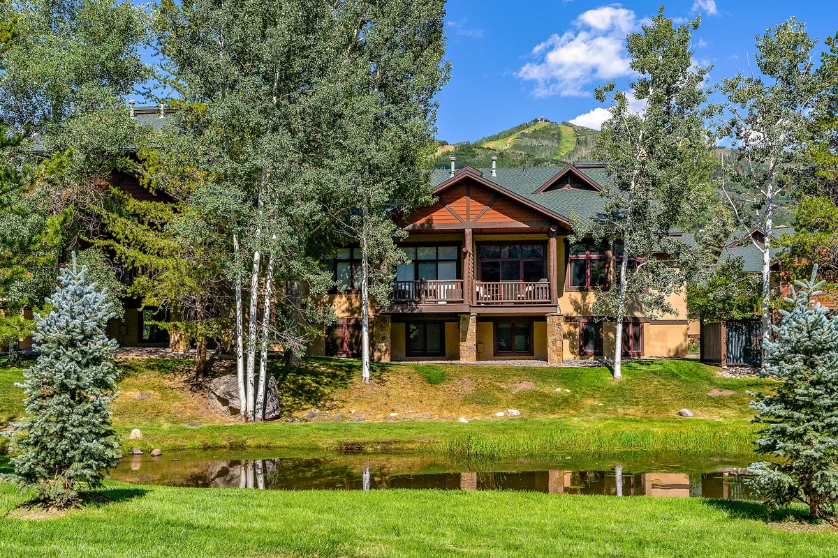 The hero image showcases the elegant end-unit townhome nestled beside a tranquil pond, framed by lush green trees and backed by majestic mountains glowing in the sunset. The spacious deck overlooks the peaceful water, with ample space for relaxing or gathering with friends and family. The setting evokes a sense of serenity, connection to nature, and retreat from daily life.